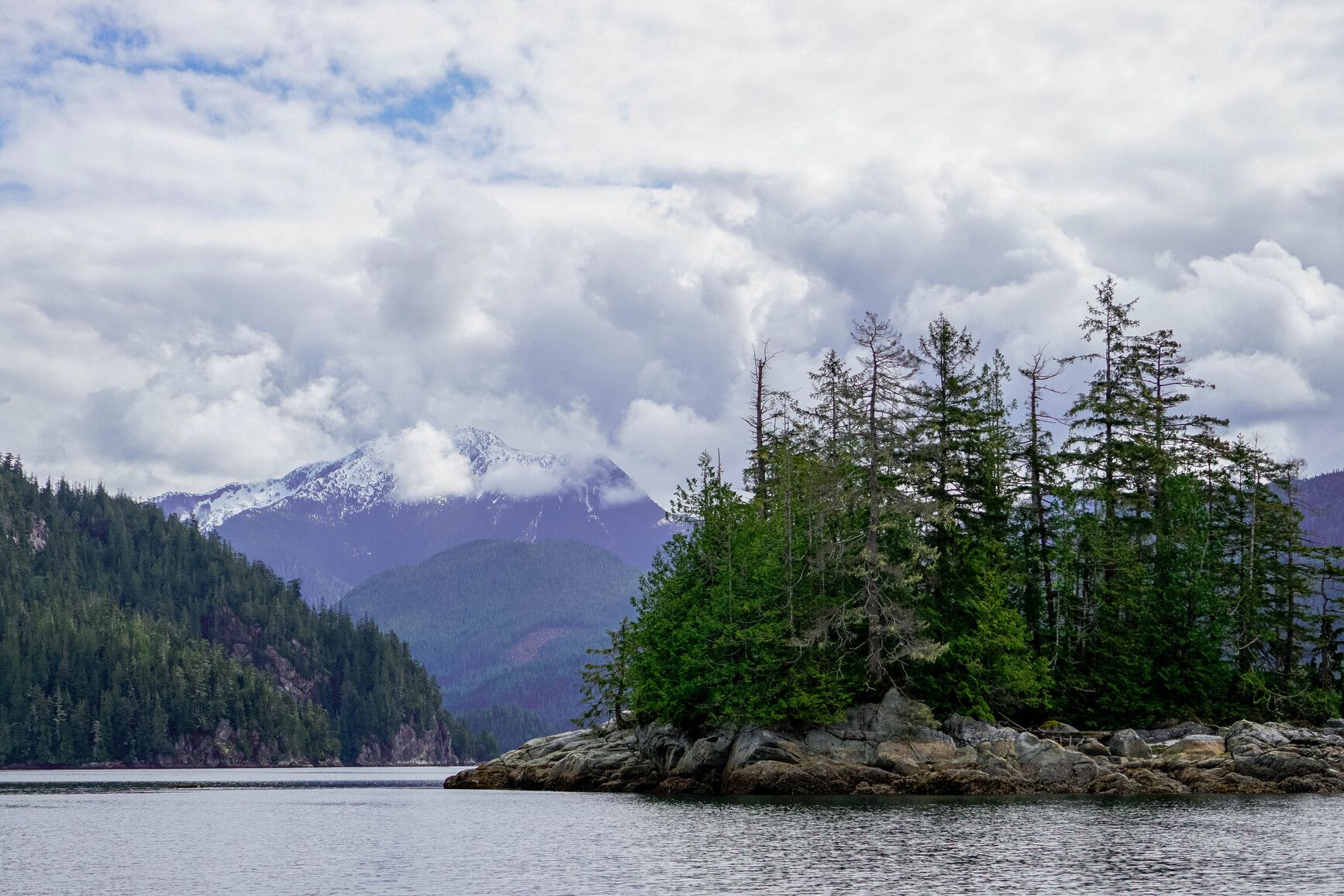 <p>A calm coastal scene where forested islets meet quiet water, backed by mist and distant mountain peaks in Broughton Archipelago Park.</p>