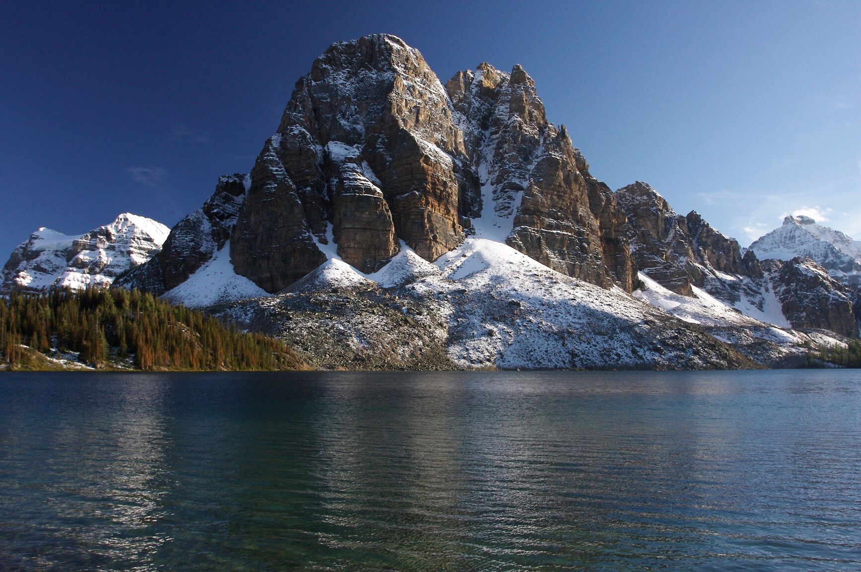 Sunburst Peak rises above the clear waters of Cerulean Lake in Mount Assiniboine Park, framed by fresh snow and autumn forest.