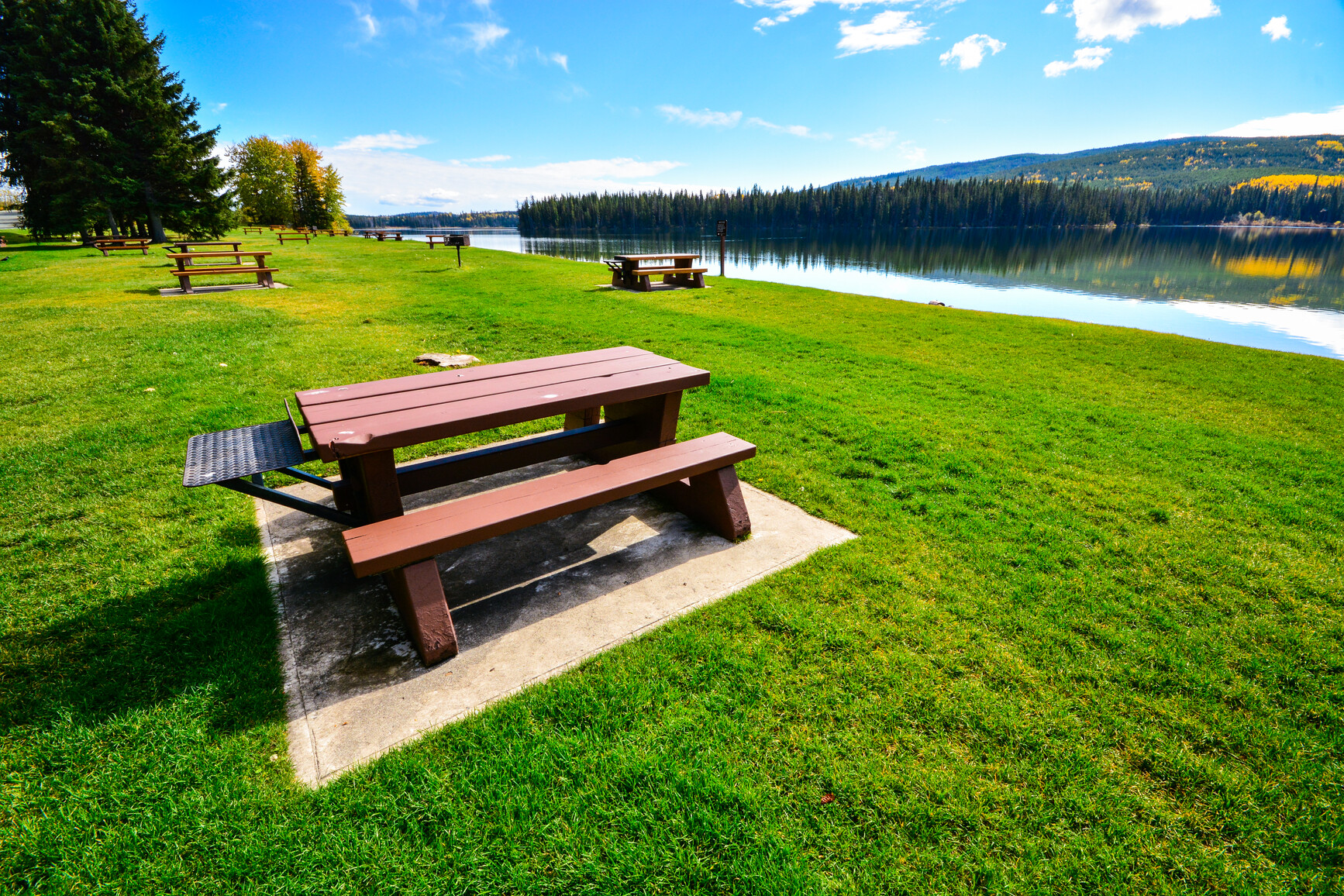 Picnic area of park. Several picnic tables in foreground.  Lake and mountains in view.