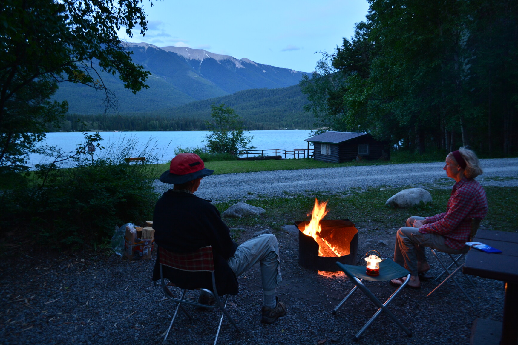 Campers sitting by fire. View of lake and mountains.