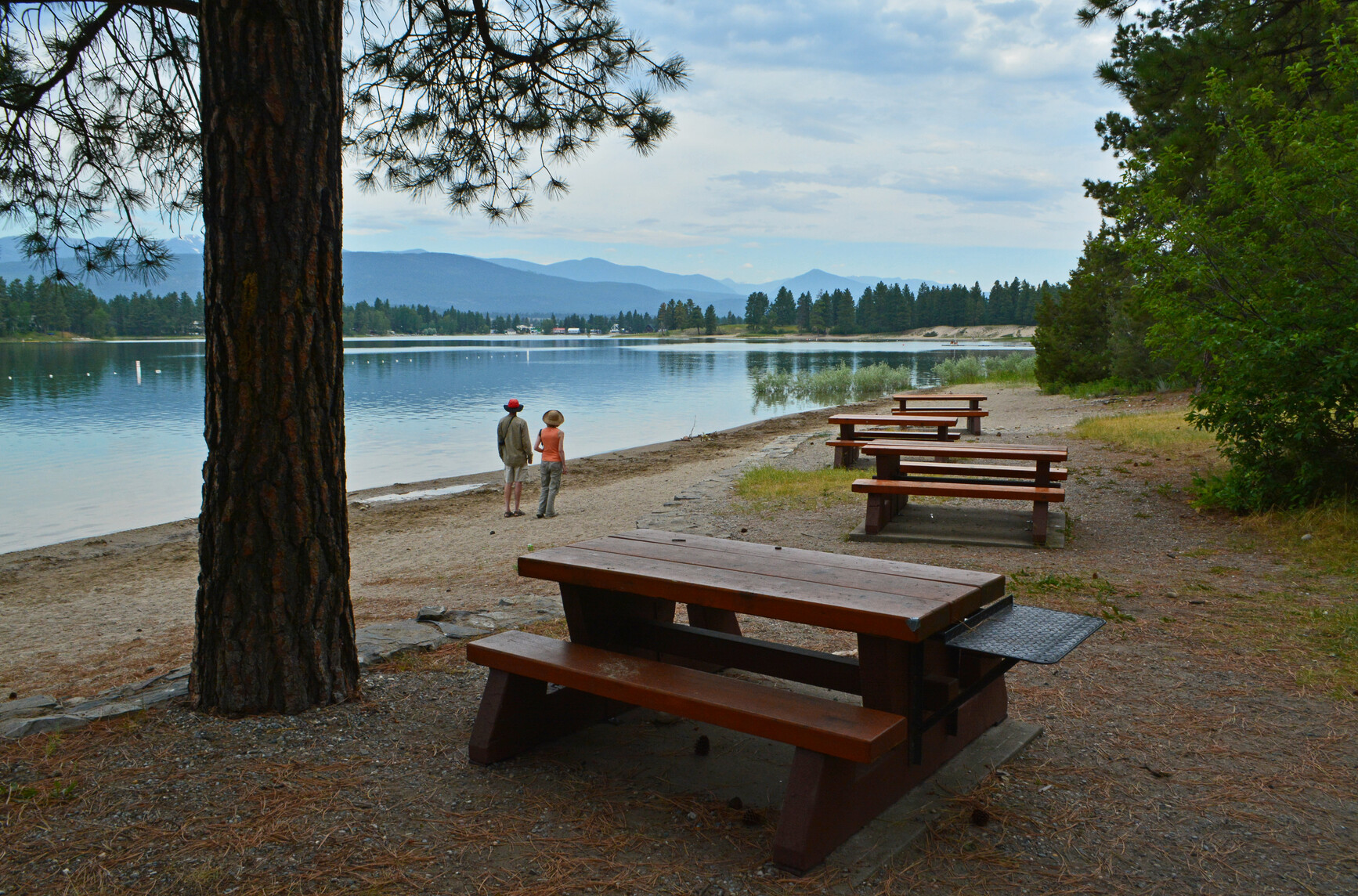 Park visitors standing on the beach beside the picnic area looking at the view.  Mountains are in view on the other side of the lake.
