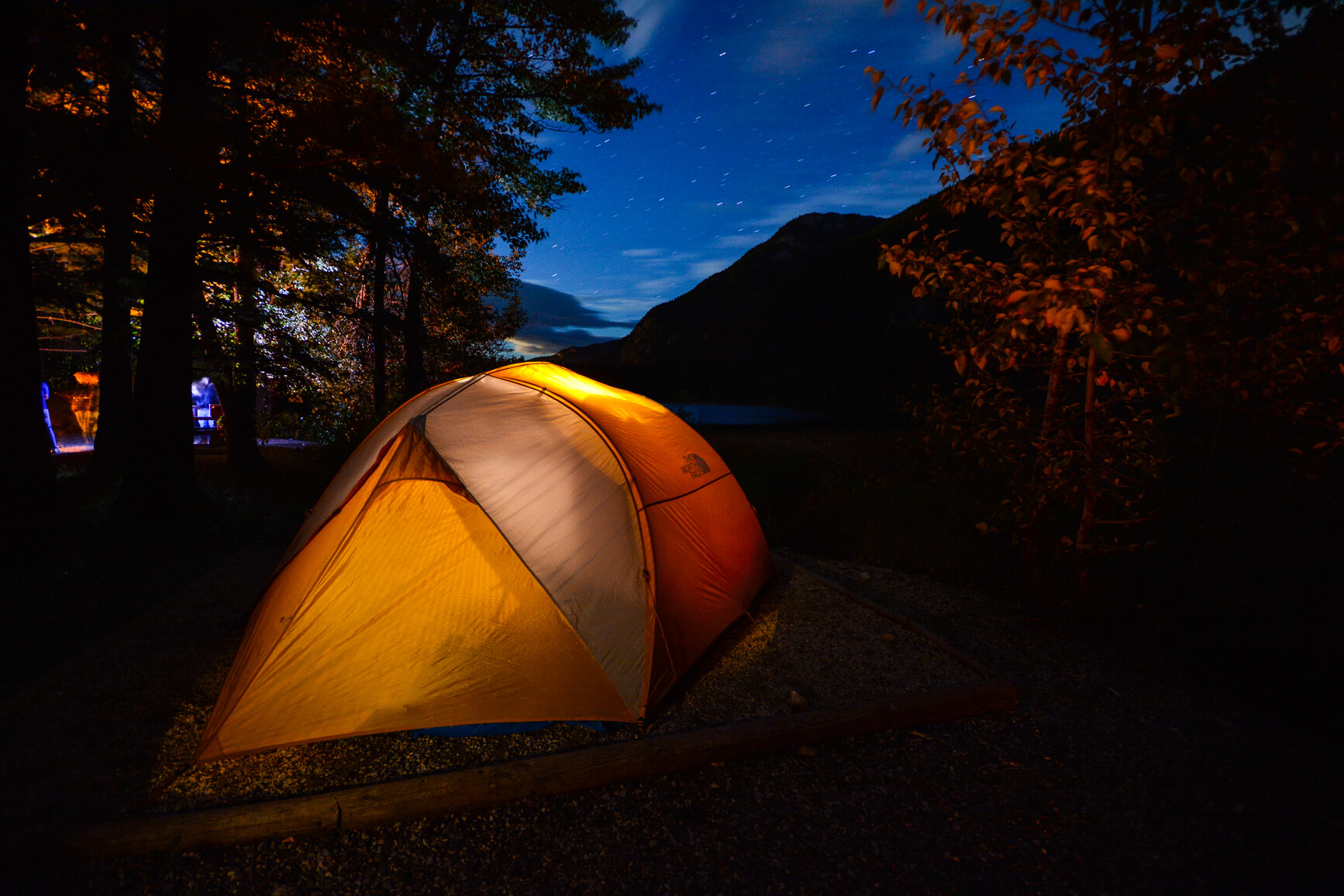 Campsite at night with a tent glowing from a light on inside. A fire is burning in the fire ring. In the background is a lake and forest covered mountains.