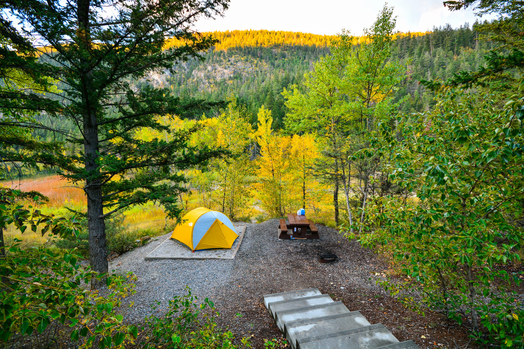 A forested campsite with stairs leading down to it, a tent on a tent pad, a picnic table, a fire ring. In the background are forest covered mountains.