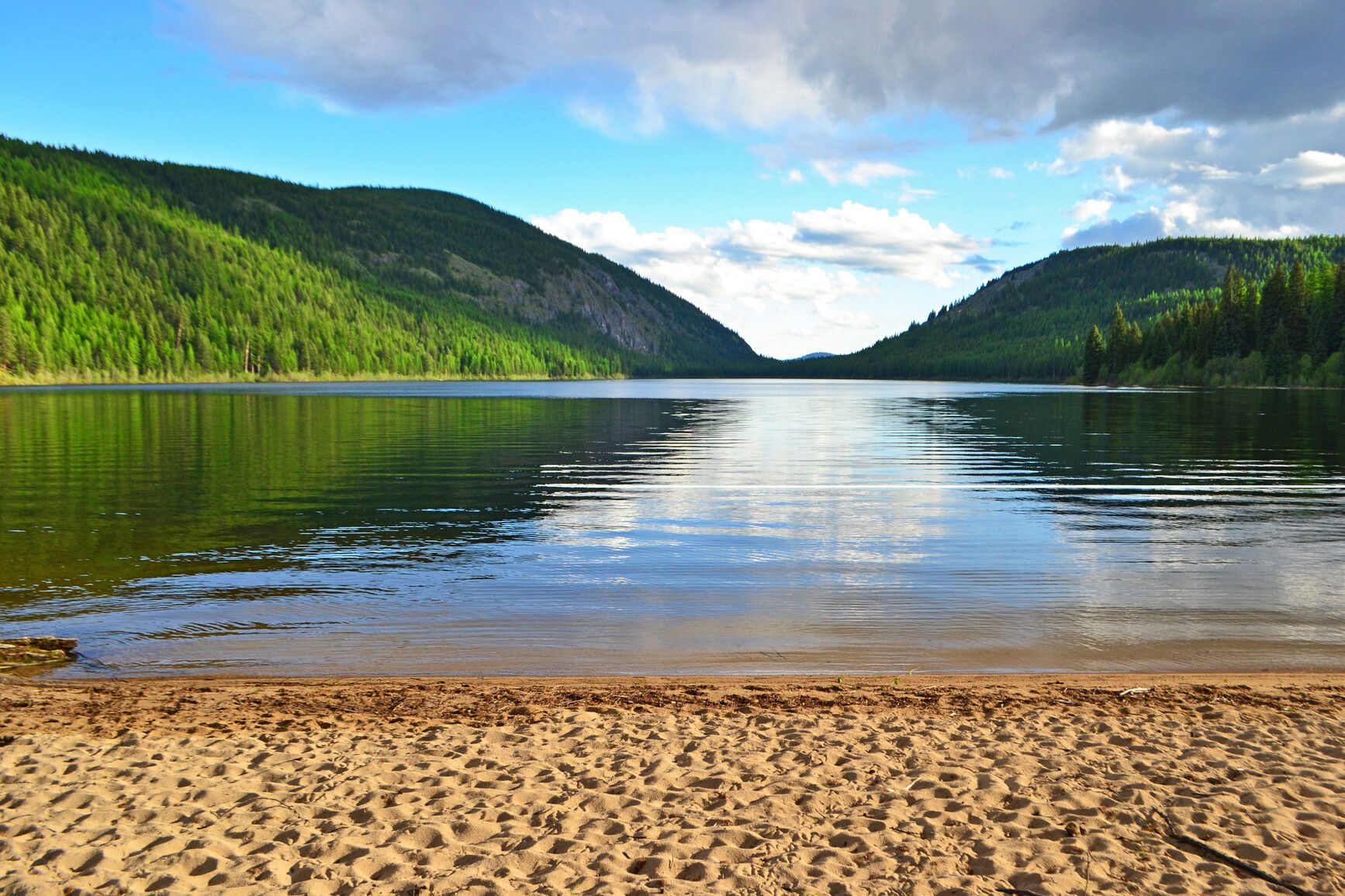 Discover the serene beauty of Conkle Lake Park with its sandy beach and breathtaking vistas of forest-covered mountains.