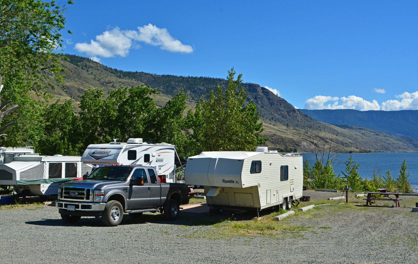 Kamloops Lake - Campground with truck, trailers, tent trailers and power hook up. Picnic tables on the right and trees, lake and mountains in the background.
