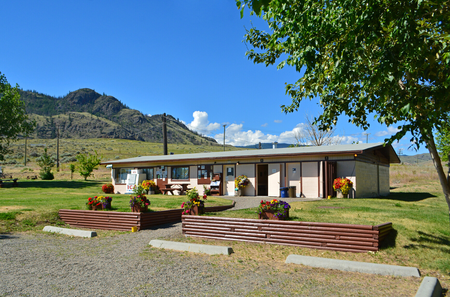 A gravel walkway leading to a grassy area and shower building. Icebox, picnic table and potted flowers are in front of building. Forested mountains and trees are in the background.