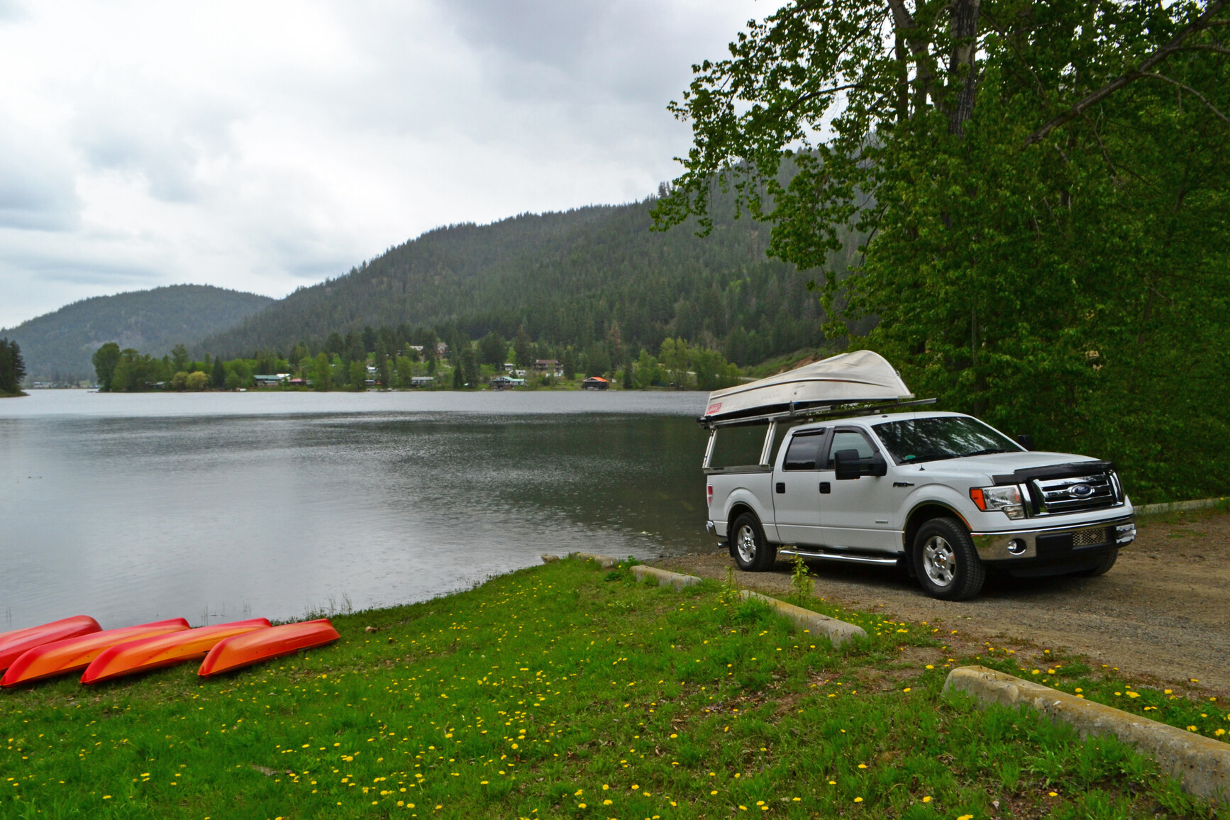 A truck preparing to launch a power boat at a boat launch on the lake.  On the grass in the foreground are 4 orange canoes. Trees, forest and mountains are in the background.