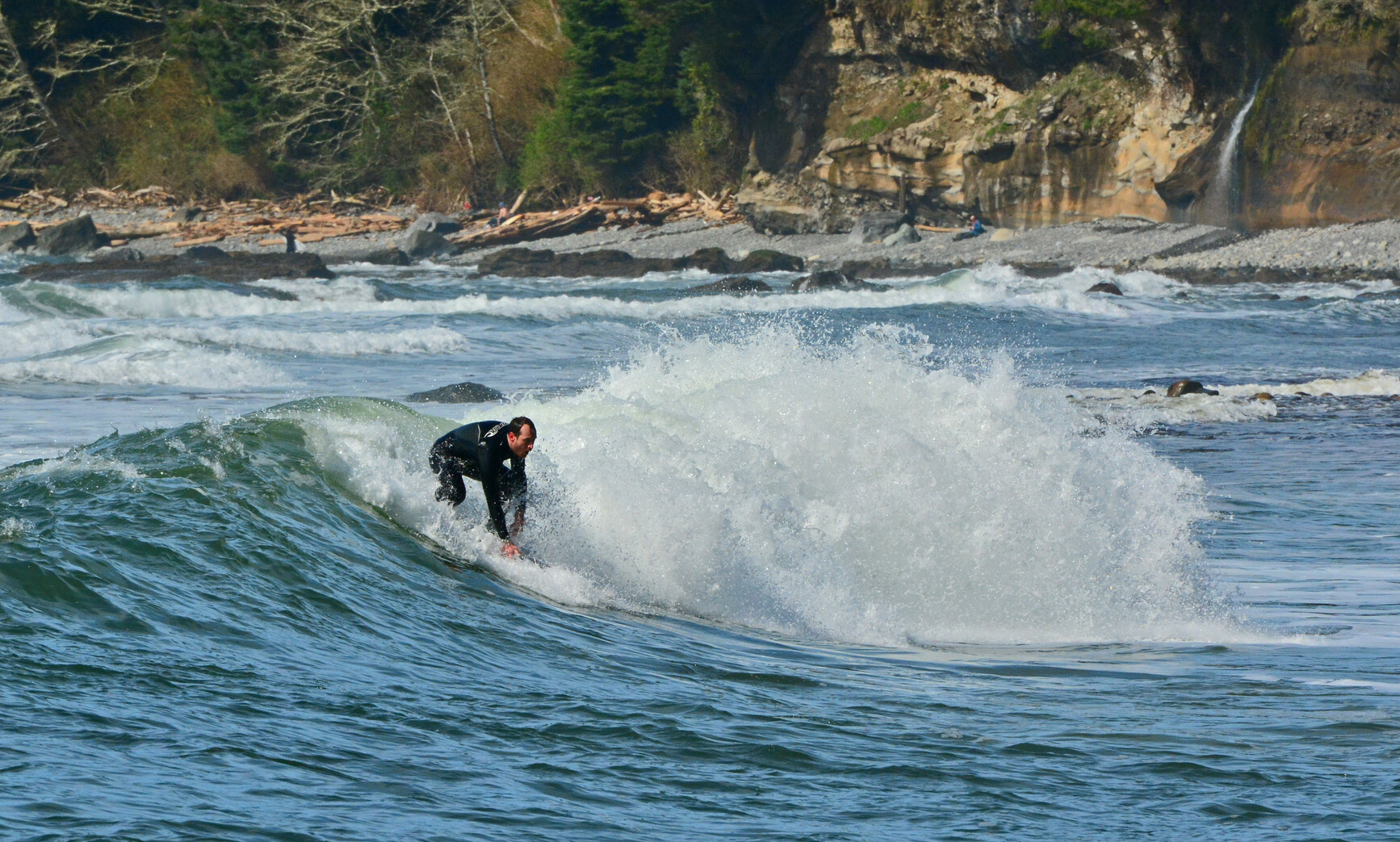 A surfer rides a wave at Sombrio Beach in Juan De Fuca Park. Along the beach, logs lay in front of the rocky and forest covered sandstone walls of the shoreline.