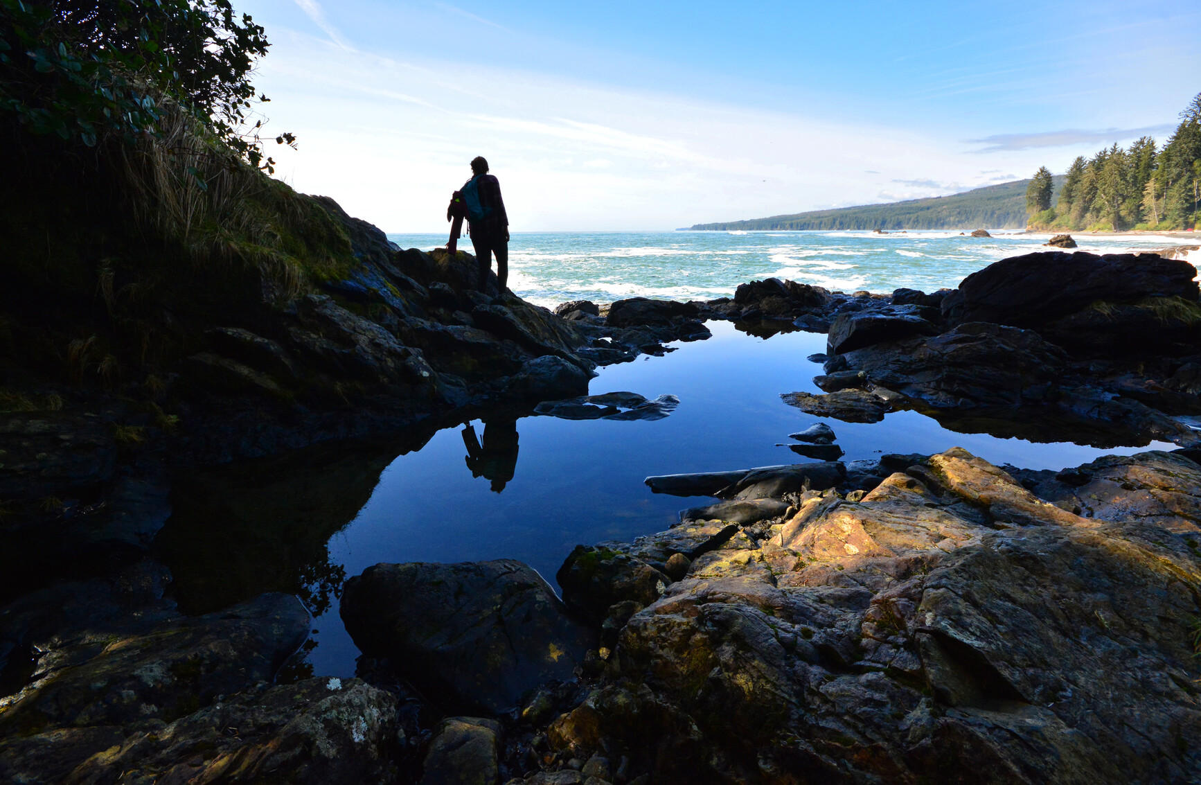 The silhouette of a hiker in the shade by a tide pool looking out at the ocean view, with the backdrop of mountains and forest. Sombrio Point in Juan De Fuca Park.