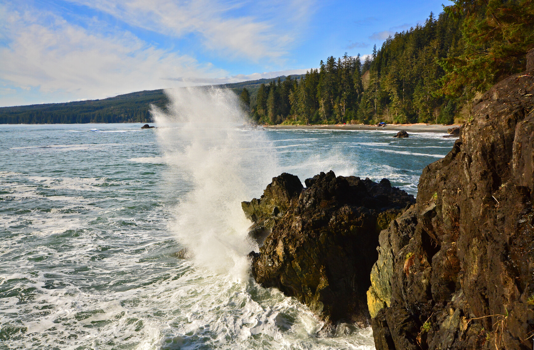 At Sombrio Point in Juan De Fuca Park, waves crash into a rocky outcrop. Along the shoreline, forest covered mountains are in view.