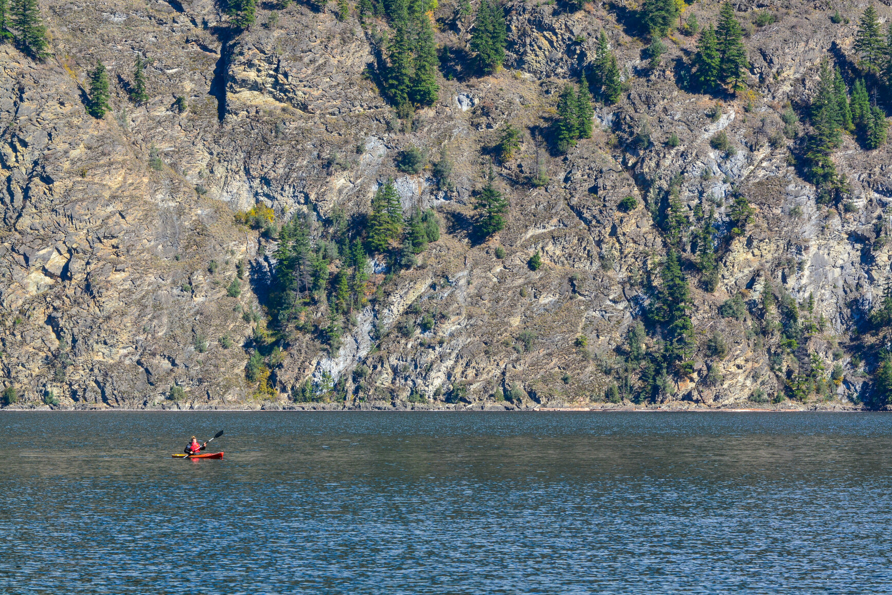 A park visitor kayaking on Adams Lake. Across the lake are rugged cliffs with a few trees.