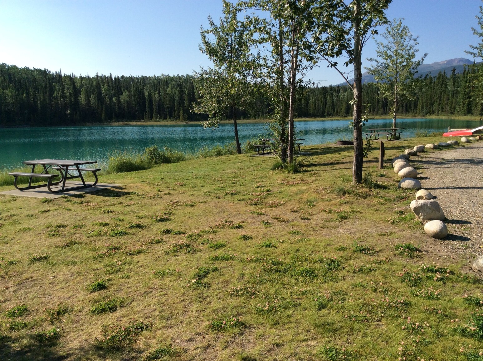 Grassy day use area with picnic tables and a few trees on the edge of the lake. Canoes on the beach in the background. Blue lake and forested mountains in the distance.