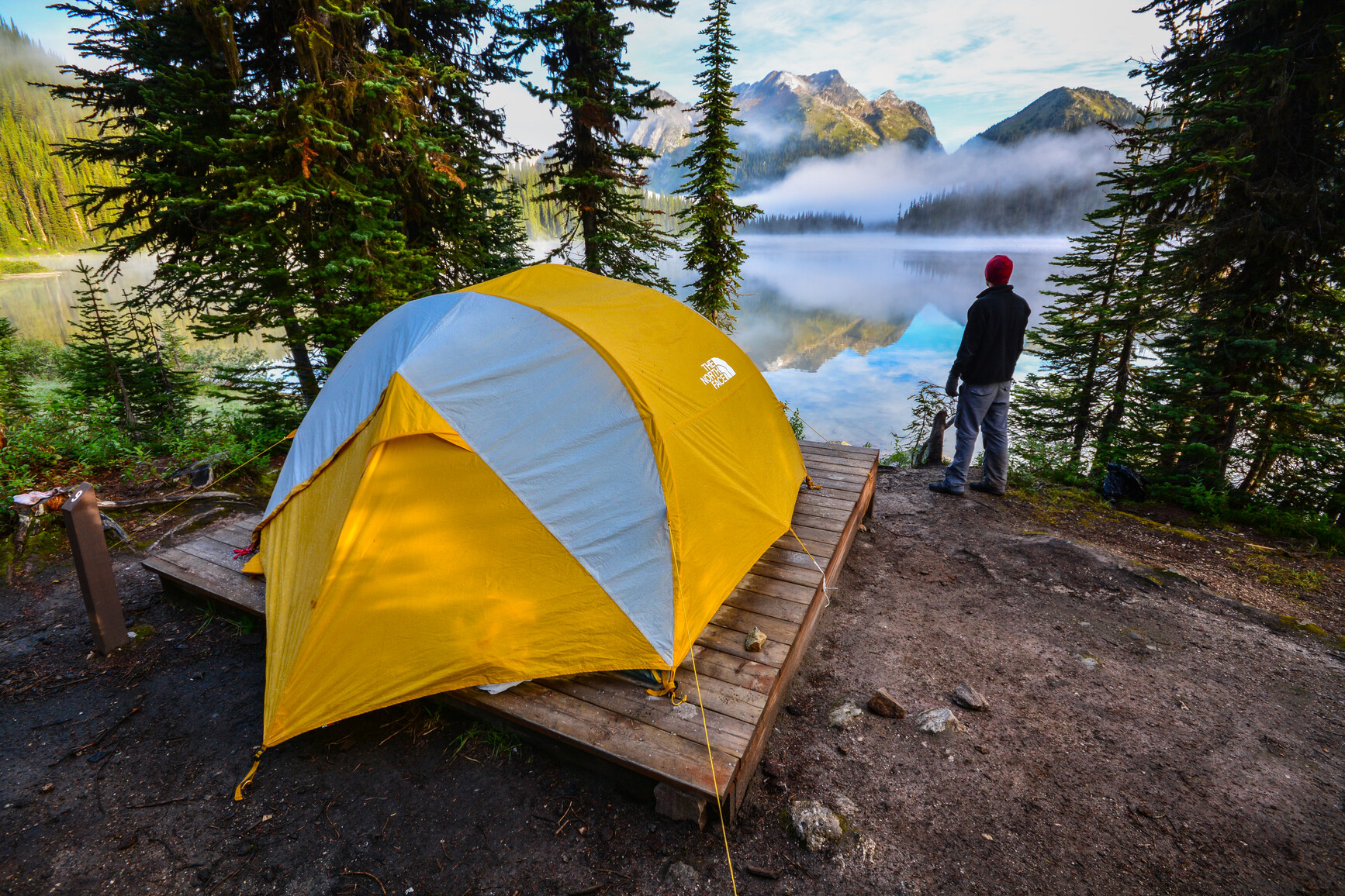 Photo of a tent on a tent pad with a view of Big Peters Lake. The campsite has sparse trees. Across the lake is forest and mountains. A park visitor is looking out toward the lake enjoying the view.