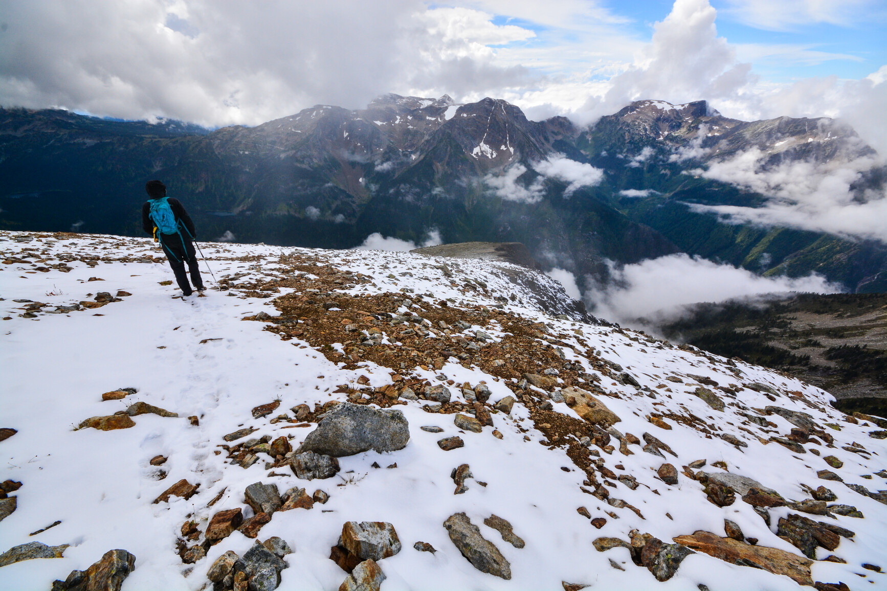 A park visitor is enjoying the view from a ridge on Mount Frosthall. The ridge is snowy and consists of rocky terrain. Across the valley is big mountains with summits in the clouds.
