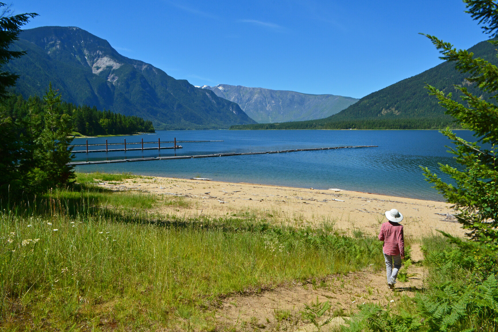 McDonald Creek Park, Upper Arrow Lake - A park visitor walking on a path towards a sandy beach at Arrow lake. Grasses and trees are in the foreground. The lake, trees, forests and mountains are in the background.