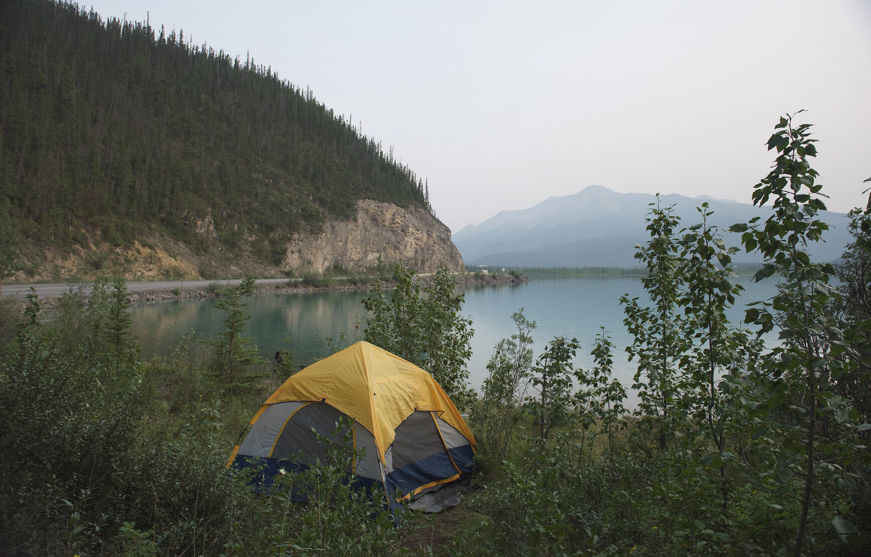 A tent set up in grass and shrubs beside Munch Lake. The road is visible winding alongside the lake. Forested mountains in the distance. Muncho Lake Park.