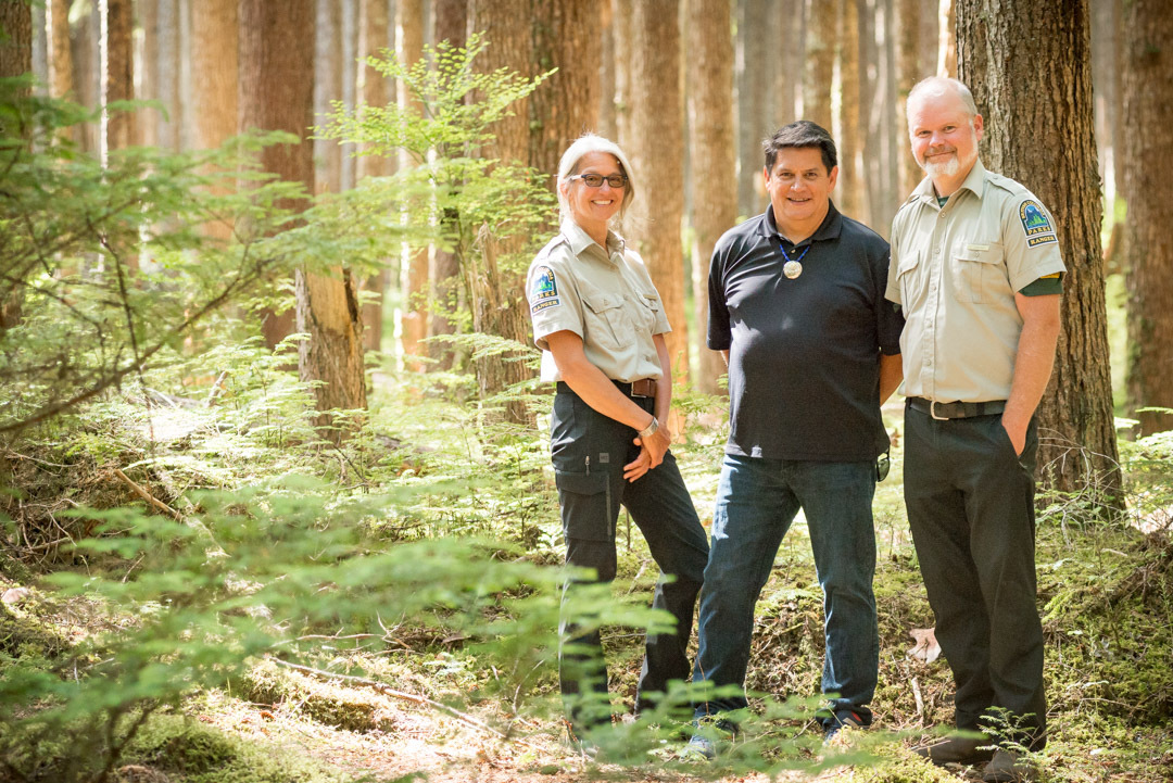 In a forested area of Naikoon Park, two park rangers stand beside Robert Russ, prior Protected Area Manager for the Council of Haida Nation.