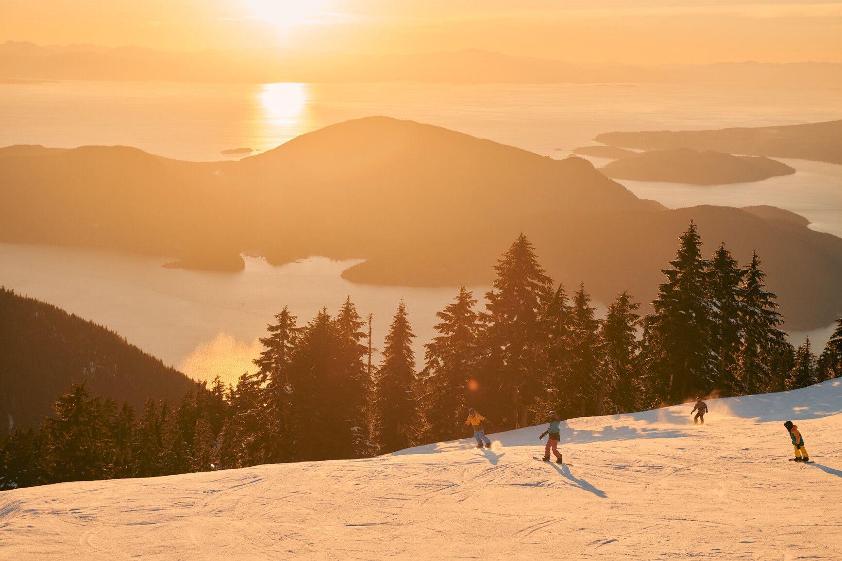 A family skiing down Cypress Mountain. The ocean and islets are lit by the setting sun. Credit: Destination Vancouver/Kindred and Scout