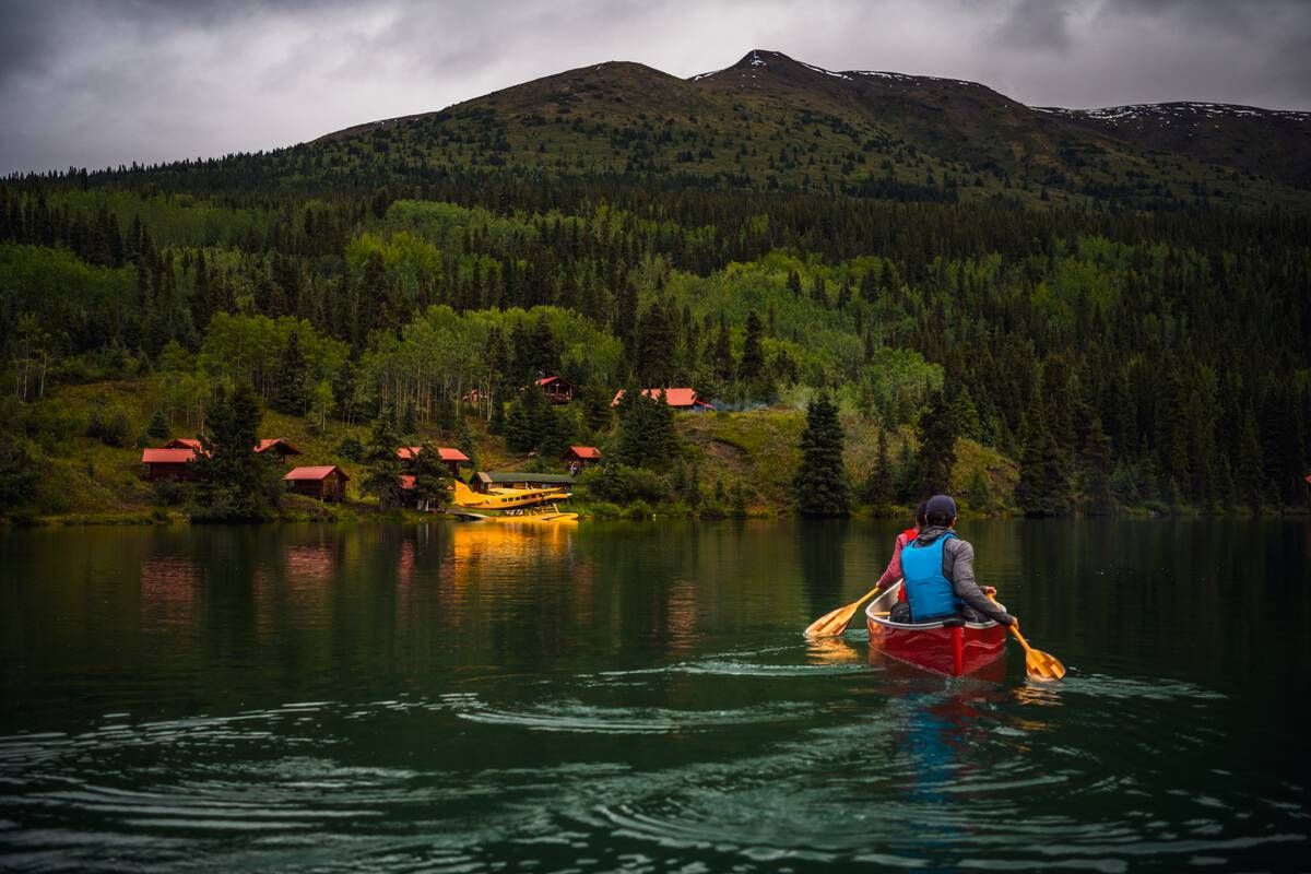 A paddler glides across Cold Fish Lake toward red-roofed cabins and a docked seaplane, surrounded by forested hills and the quiet beauty of Spatsizi Plateau Wilderness Park. Courtesy of Destination BC