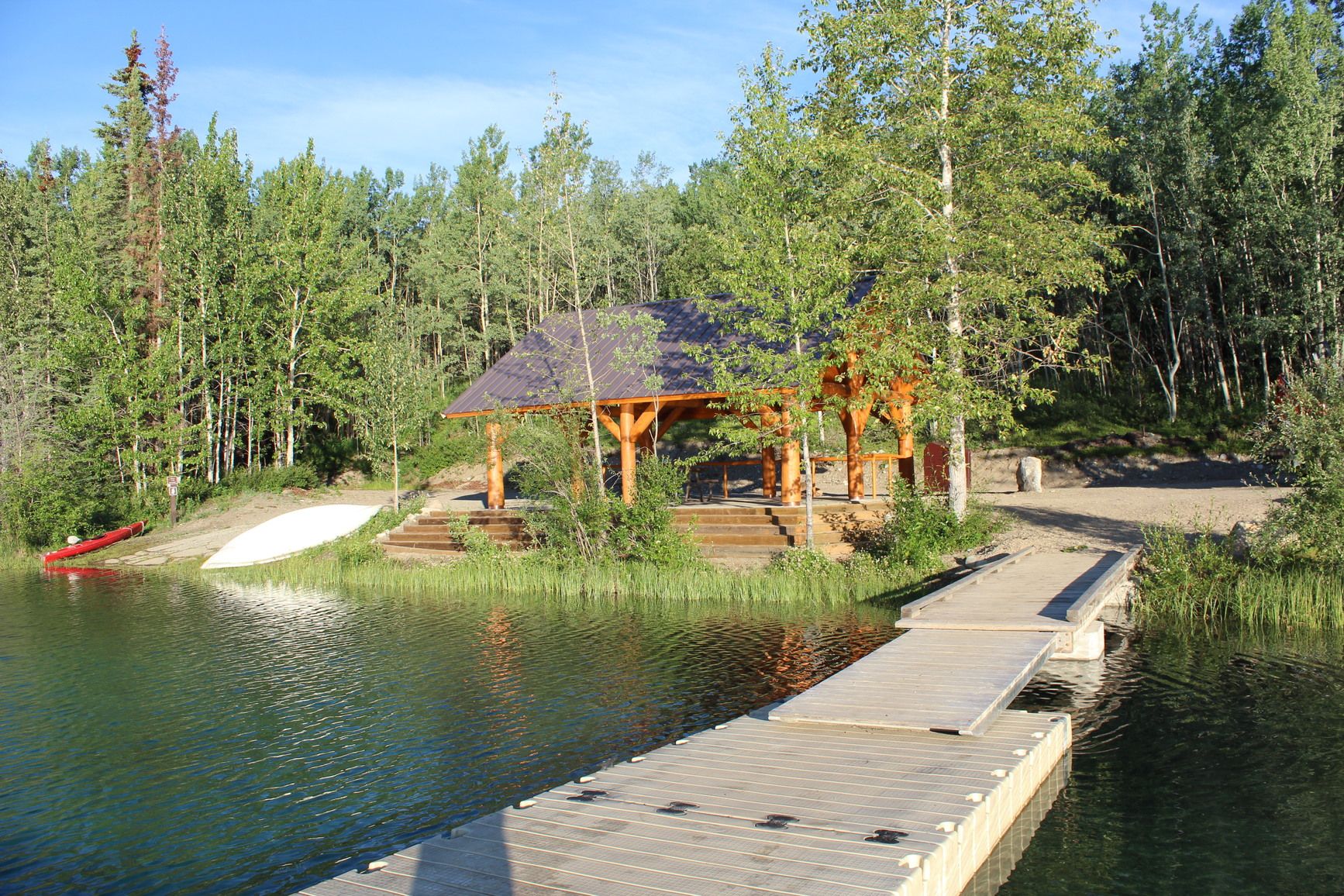 Boya Lake boat launch in Tā Ch’ilā Park features a wooden dock, a peaceful picnic shelter, and clear turquoise water surrounded by forest. It is an ideal spot for paddling, relaxing, and enjoying the lakeside view.