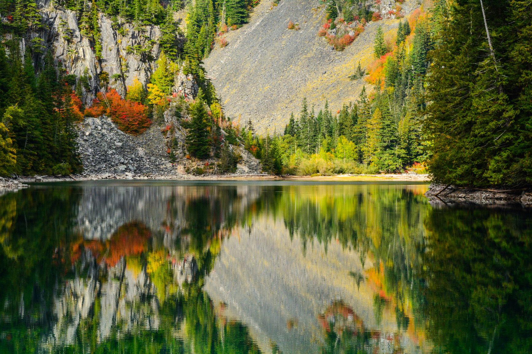 The impressive rock-slide area of Lindeman Lake, with autumn trees reflecting on the surface, in Sx̱ótsaqel/Chilliwack Lake Park.