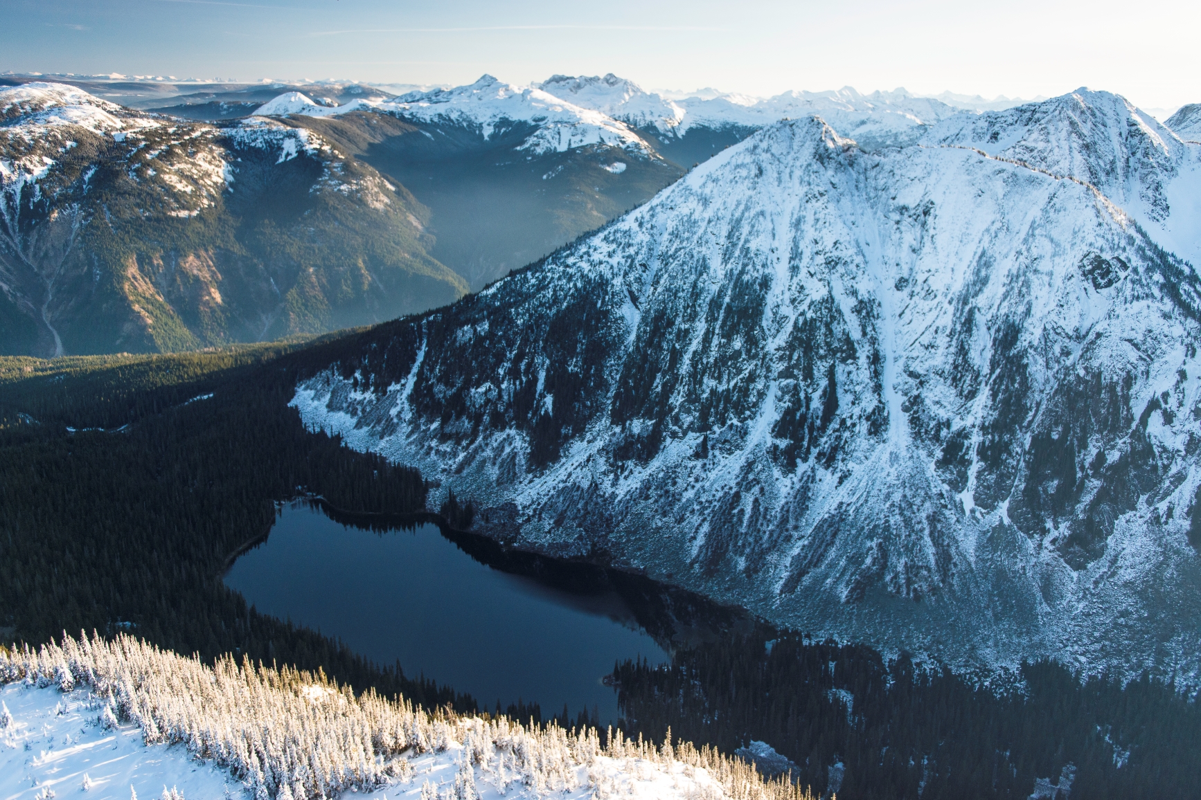 Aerial shot of the snow covered mountains, with the lake and forest in the distance.