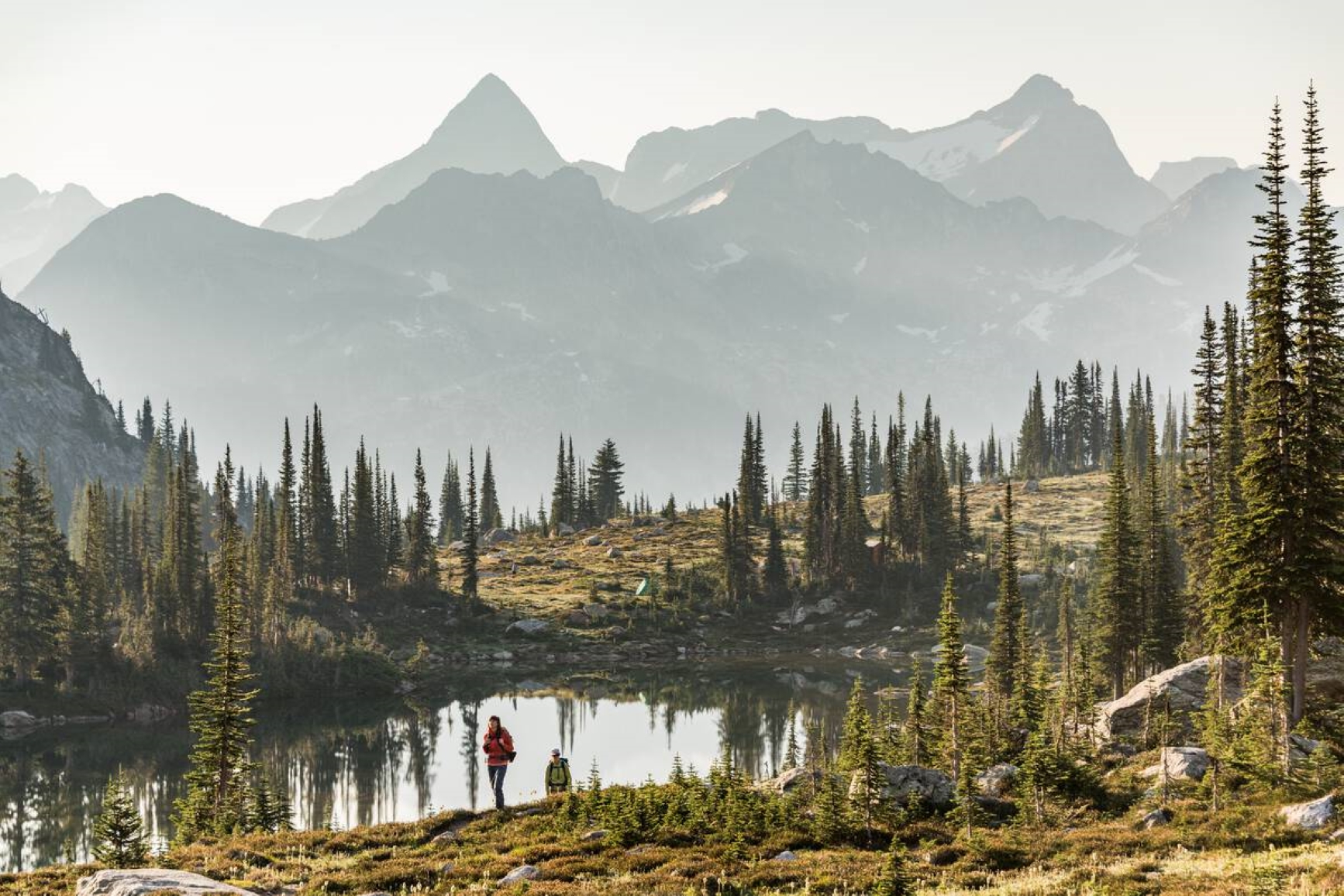 Two park visitors are exploring their surroundings. They are near a small lake that is surrounded by the forest and mountains in the distance. Photo credit: Destination BC