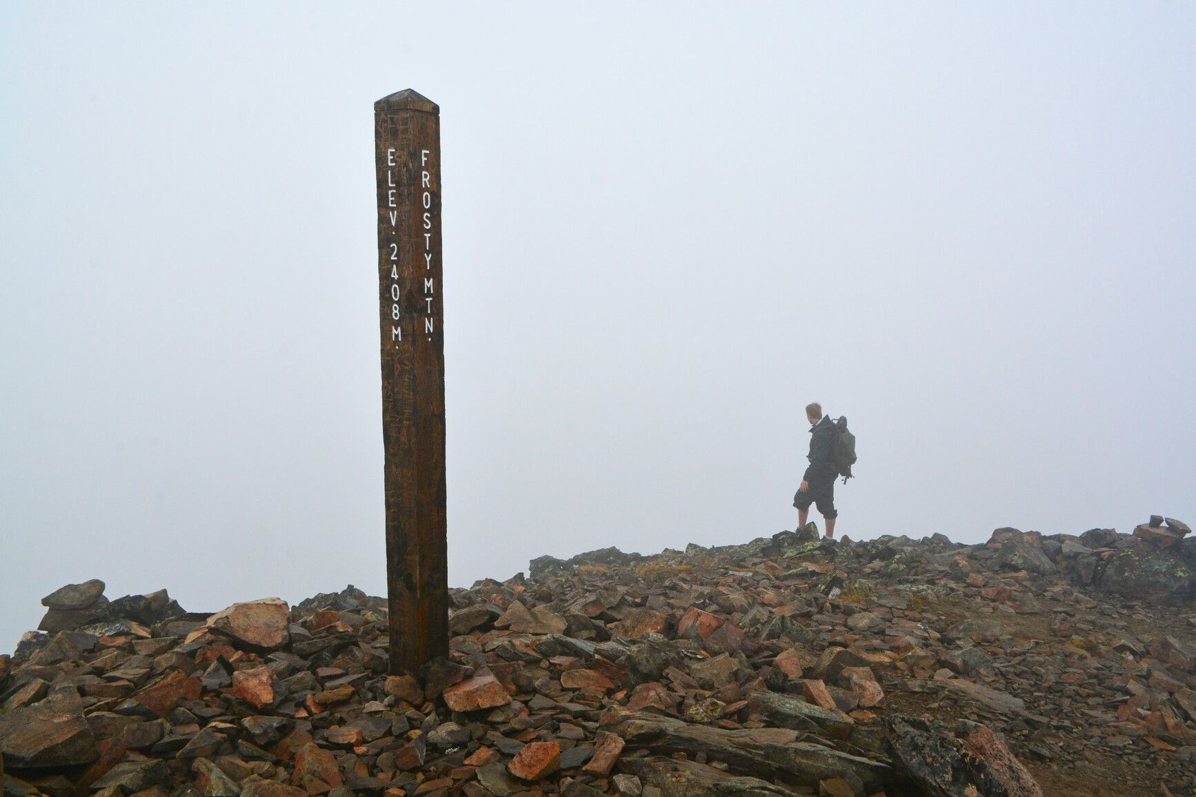 High in the alpine on Mount Frosty Trail, E.C. Manning Park, a hiker walks along a cloud-covered mountain ridge at the summit sign.
