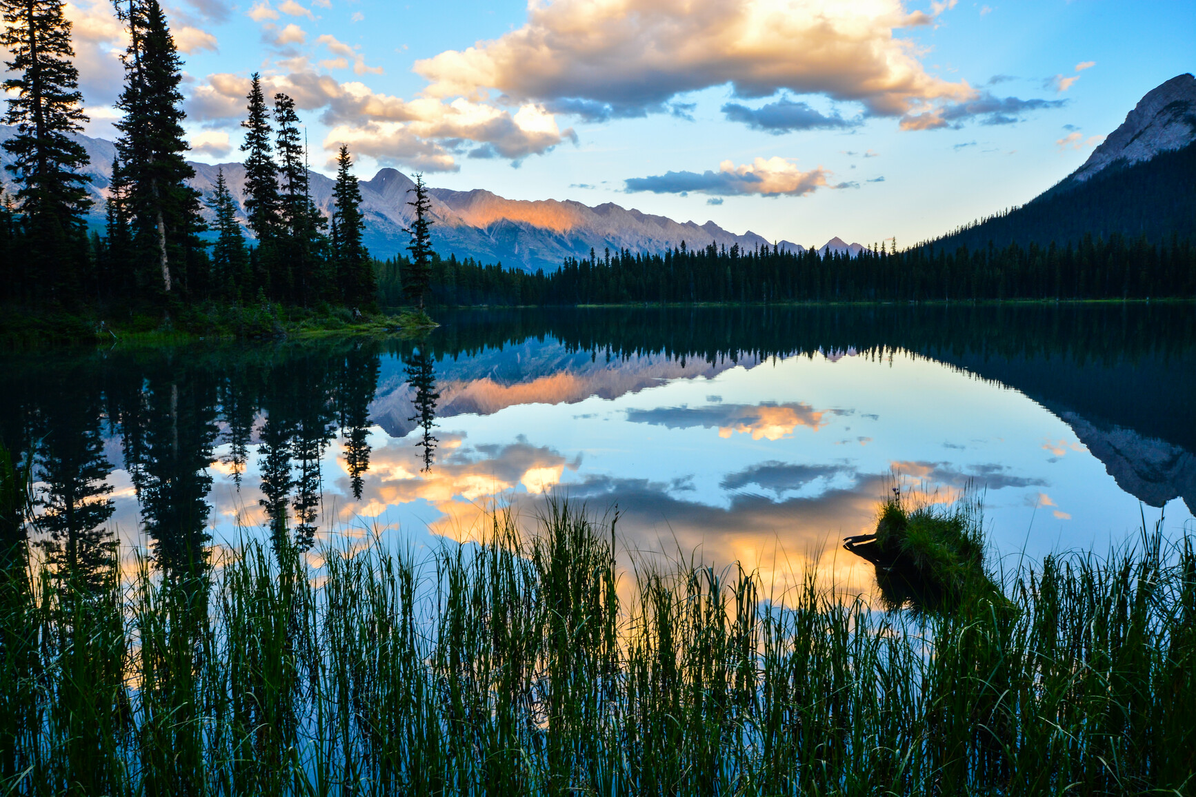 Elk Lake at early dawn. The calm lake reflects a perfect image of the surrounding forest and clouds above. The mountains glow orange in the distance.