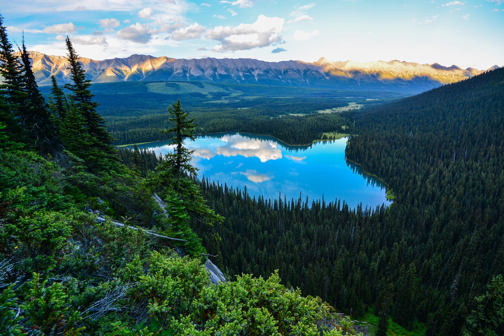 A view of Lower Elk Lake and the forested mountain valley from a slope above. The sun casts a golden glow on the mountains in the distance.