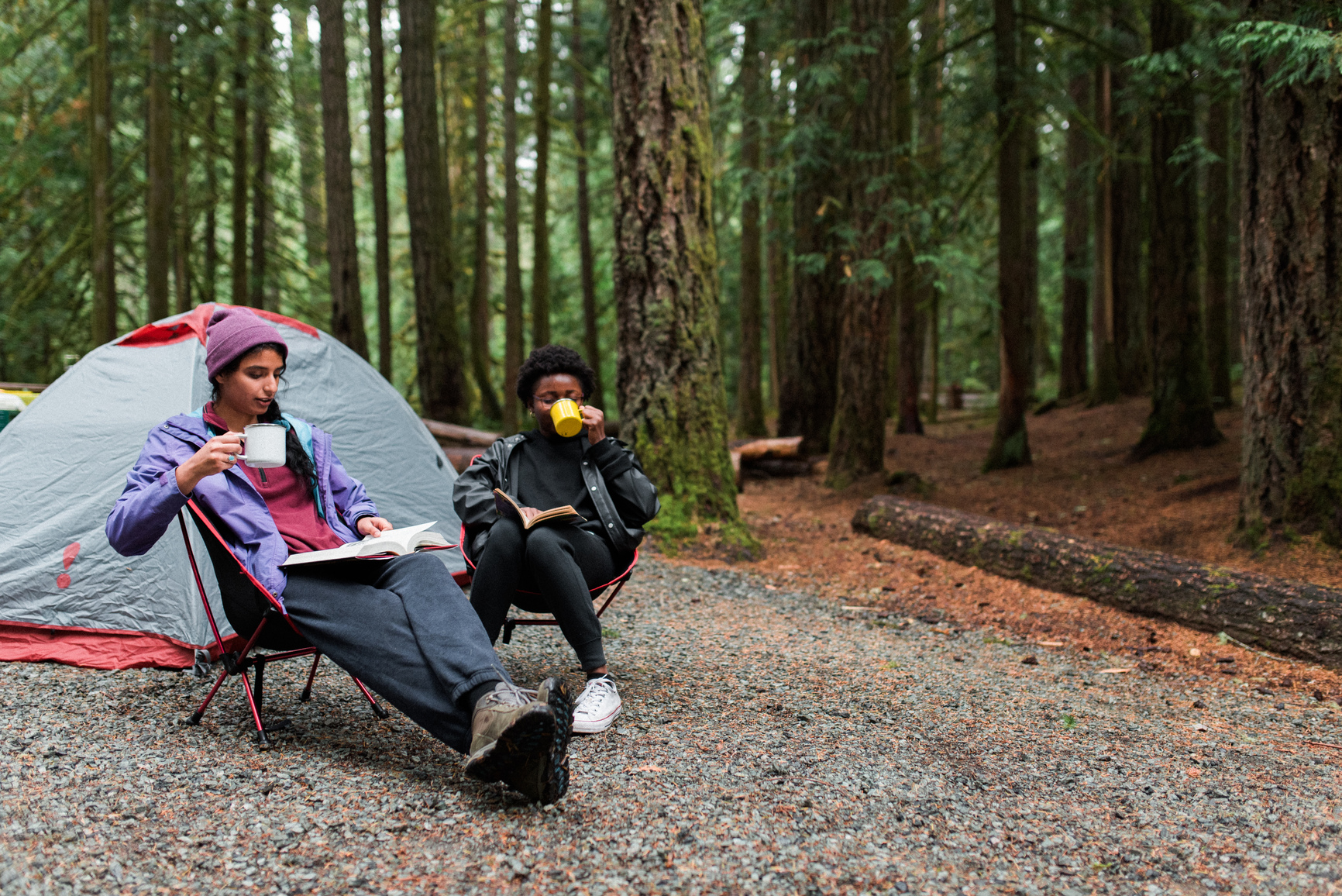 Two visitors sit in camping chairs in front of their tent, drinking coffee and reading
