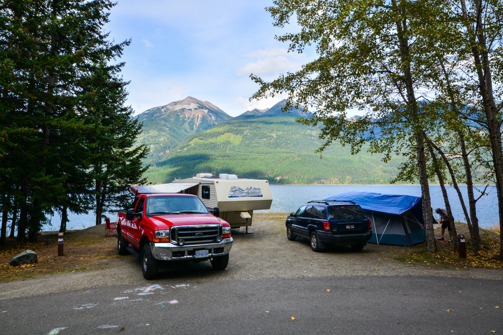 A lakeside campsite with a truck, trailer and a tent. A camper is setting up a tent. There is a view of the mountains across the lake.