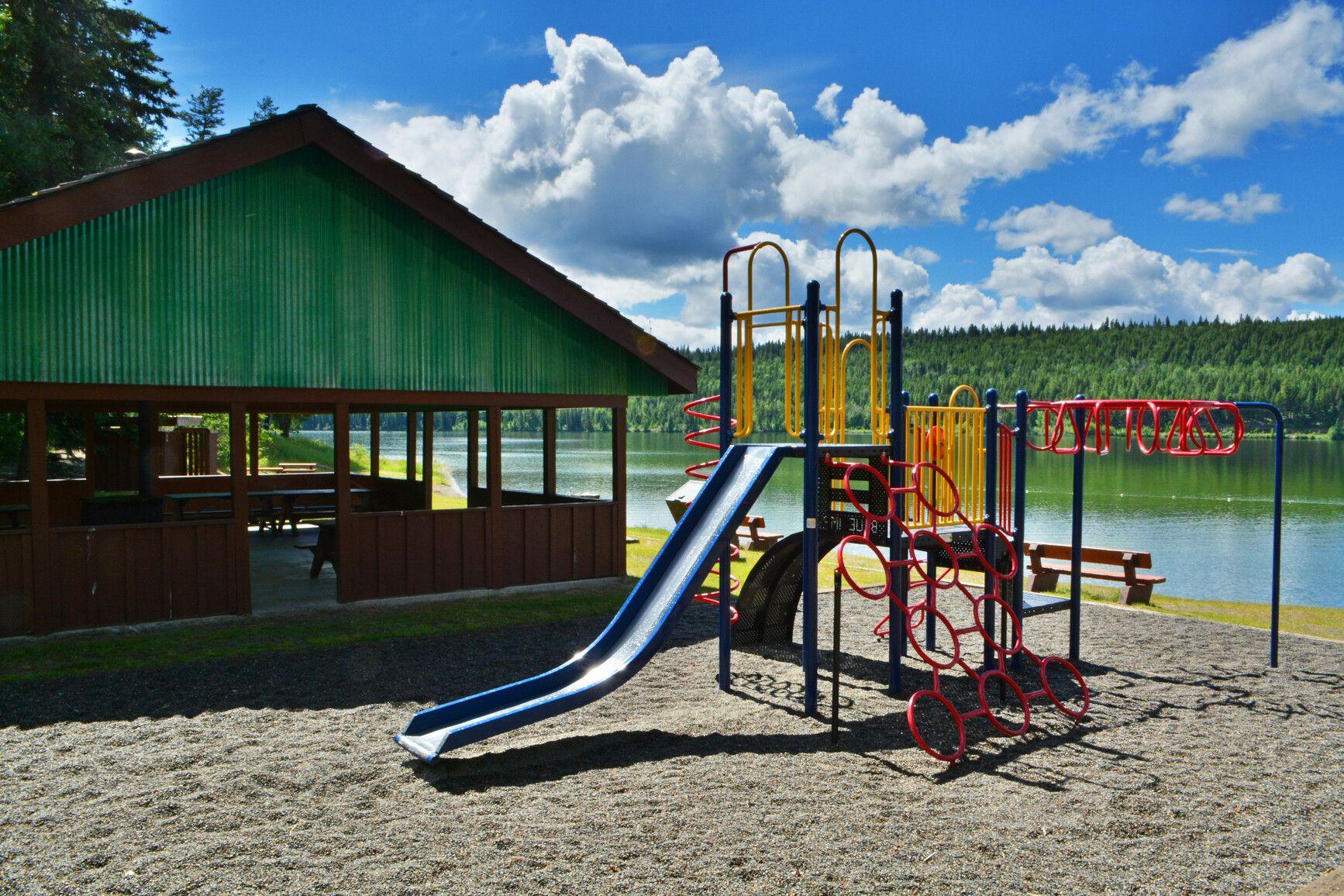 Near Lac La Hache's shoreline, you'll find a playground and picnic shelter, while across the lake lies a forested landscape.