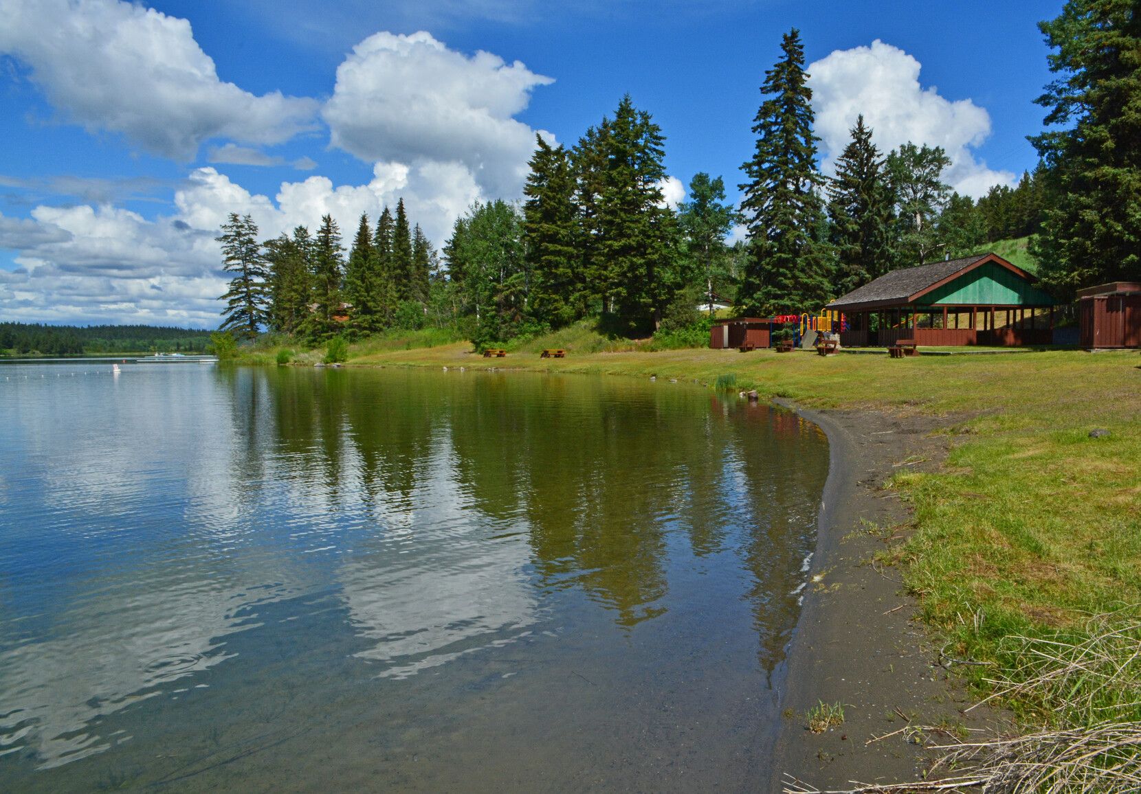Day-use area of Lac La Hache Park includes a picnic shelter, tables by the shore, and a playground.