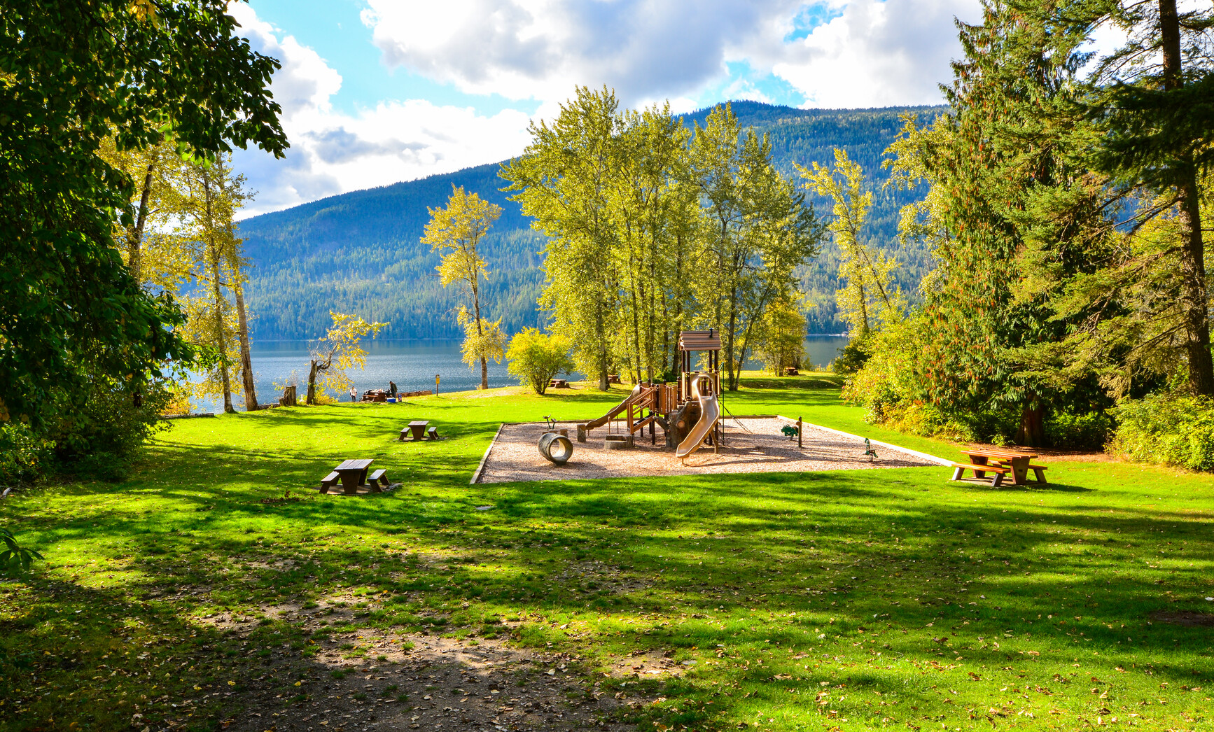 A playground at Mabel Lake Park in the picnic area. Across the lake are forest covered mountains.