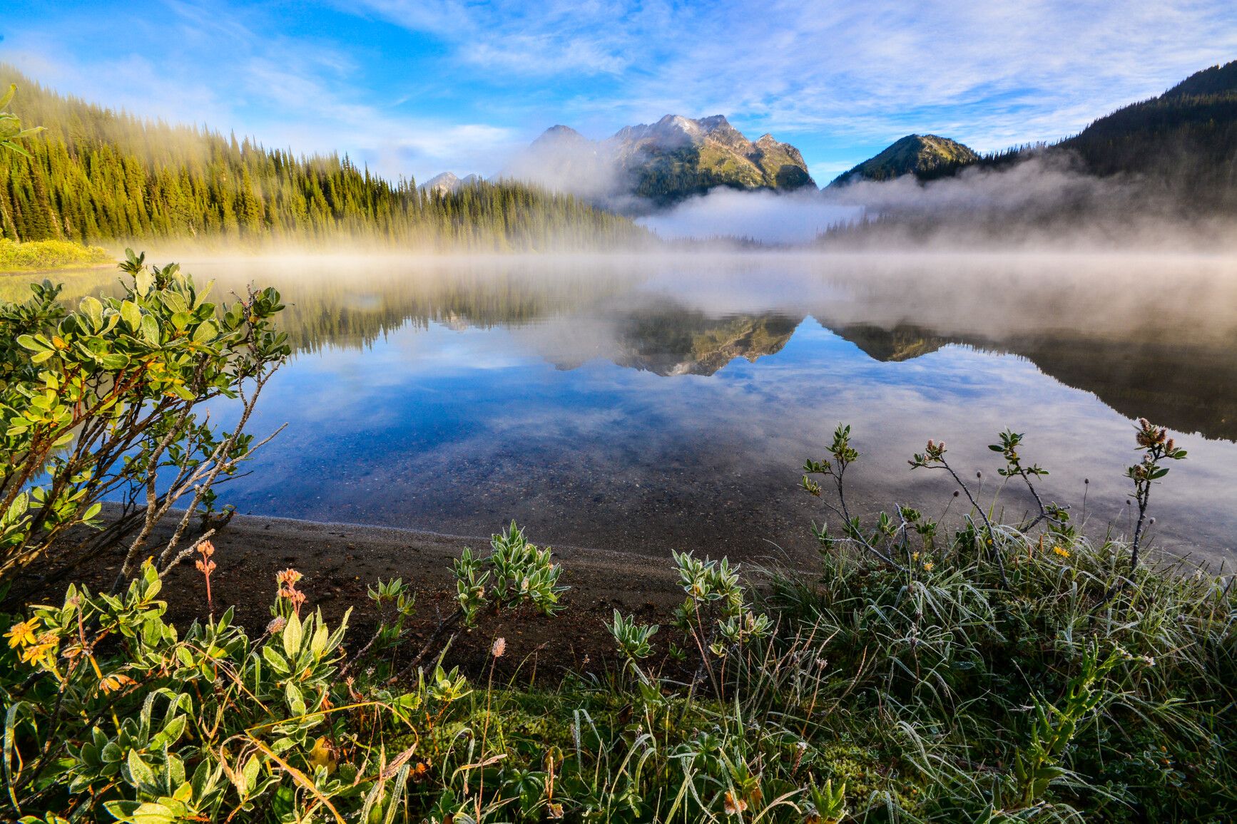 Fog rises from Big Peters Lake while the blue sky reflects on its peaceful surface in Monashee Park.