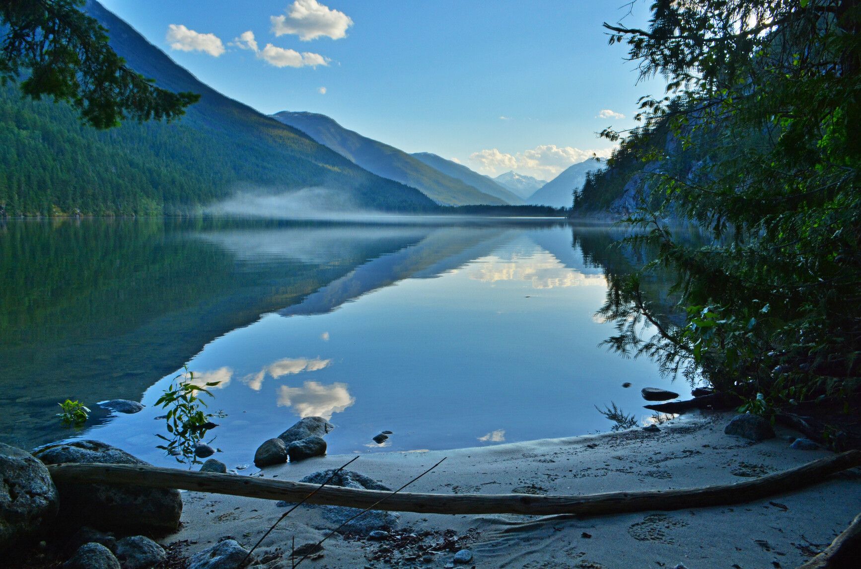Forests, mountains, and blue sky reflect on the still water of Nahatlatch Lake at sunset.