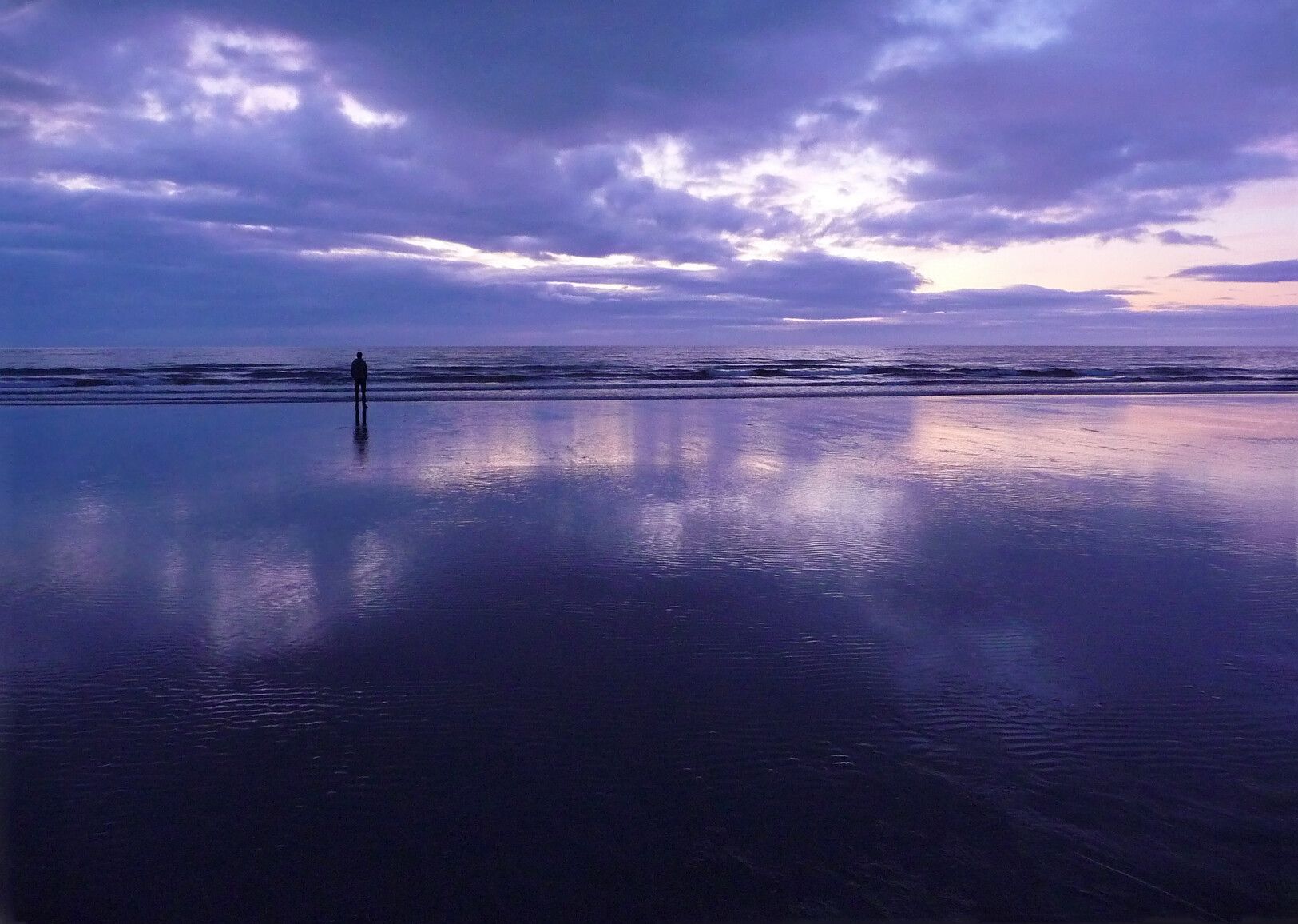 At Agate Beach, Naikoon Park, a silhouetted figure strolls along the shoreline at sunset, while the wet sand mirrors the vibrant evening sky.