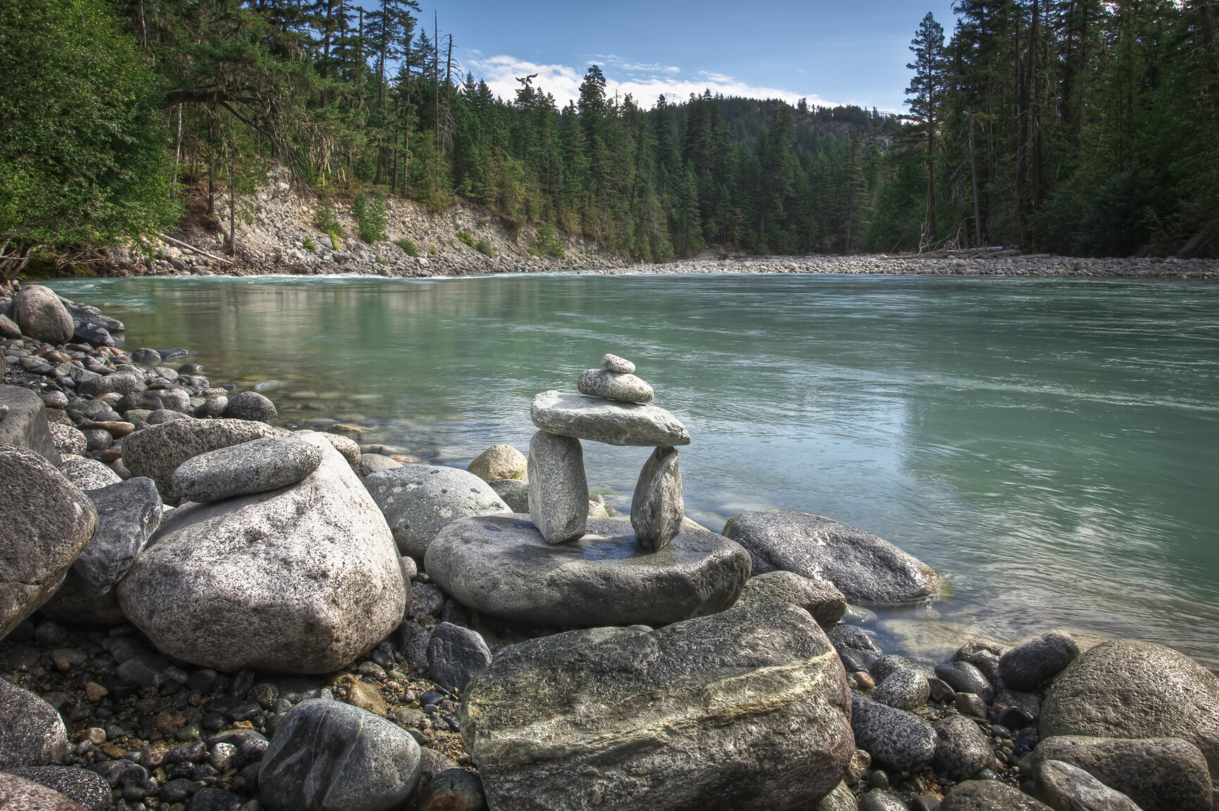 Inukshuk on rocky bank of Green River. Forest can be seen along banks of river.