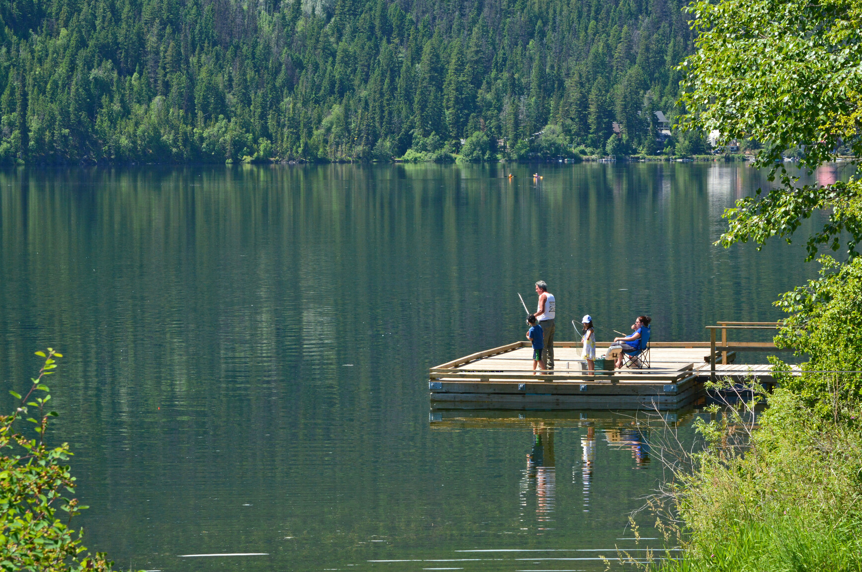 Family fishing off the dock. Kayakers on the lake. Mountains and forests in background.