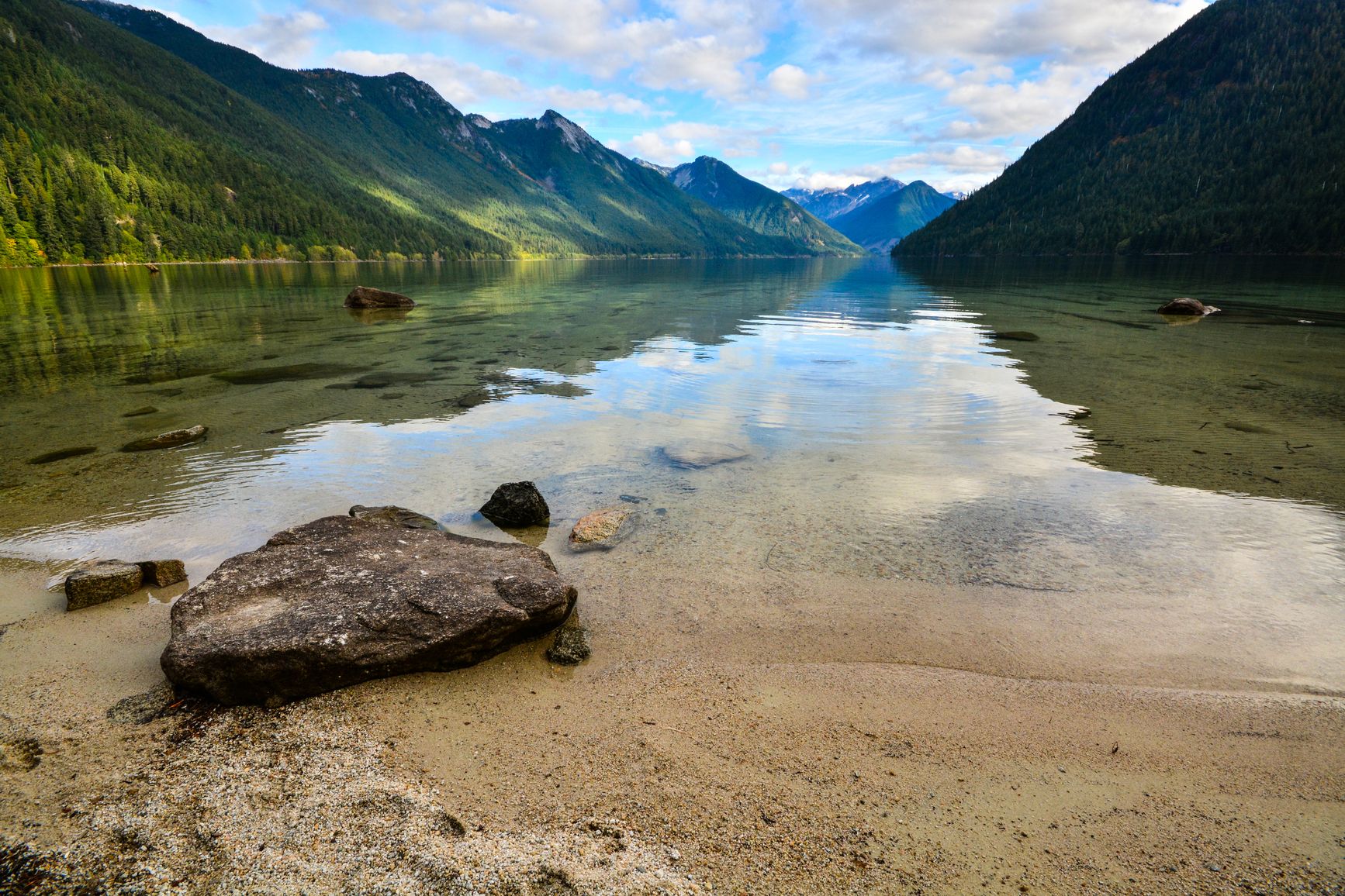 A beach in Sx̱ótsaqel/Chilliwack Lake Park offers a view of forest-covered mountains surrounding the lake.